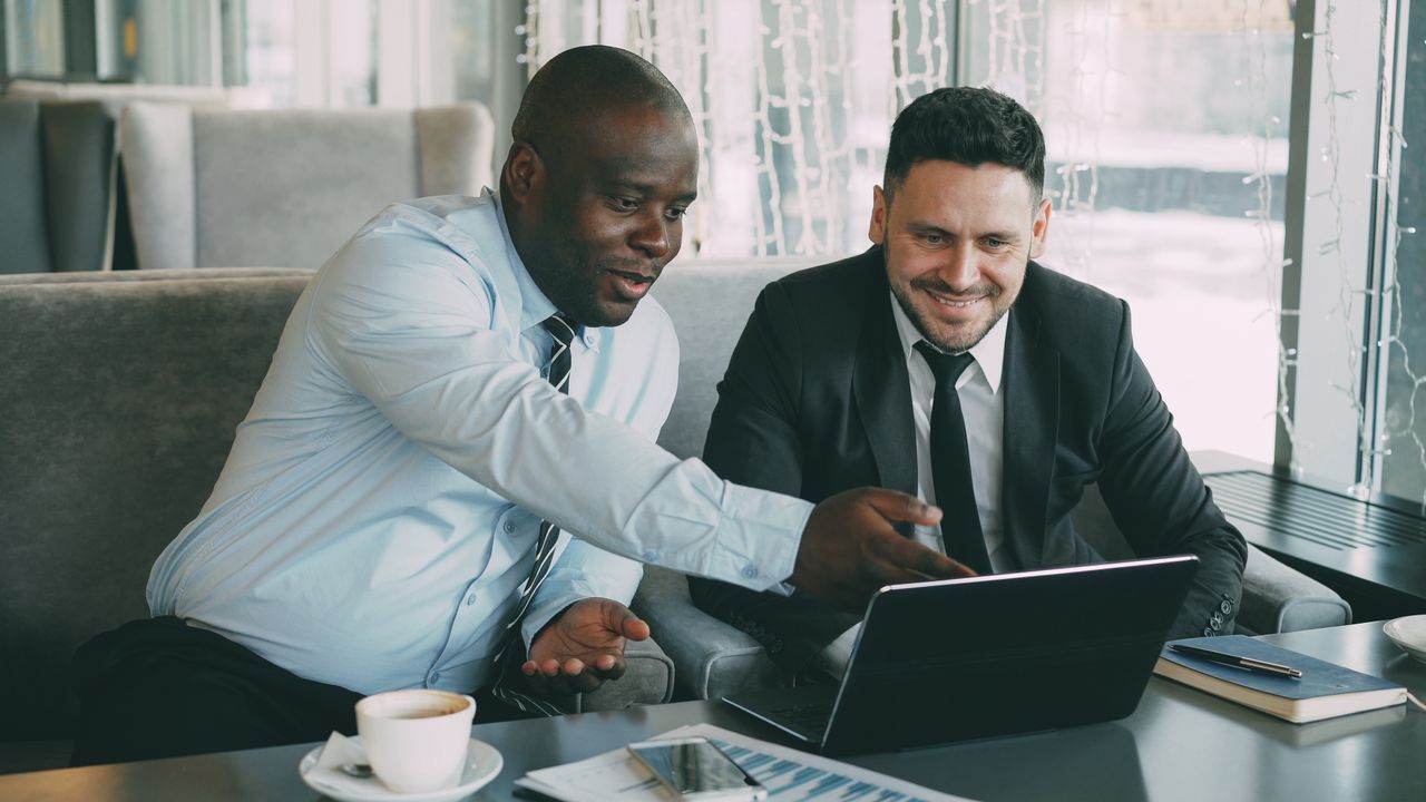 Two businessmen discussing work while using a laptop in a cafe setting.